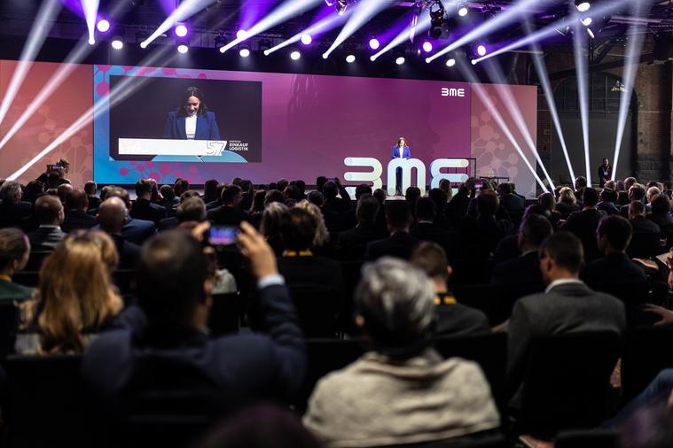 Blick aus dem Zuschauerraum auf ein Podium, auf dem eine weibliche Person spricht. Ihr Bild wird auf eine Leinwand im Hintergrund übertragen, sie selbst steht hinter einem BME-Logo.